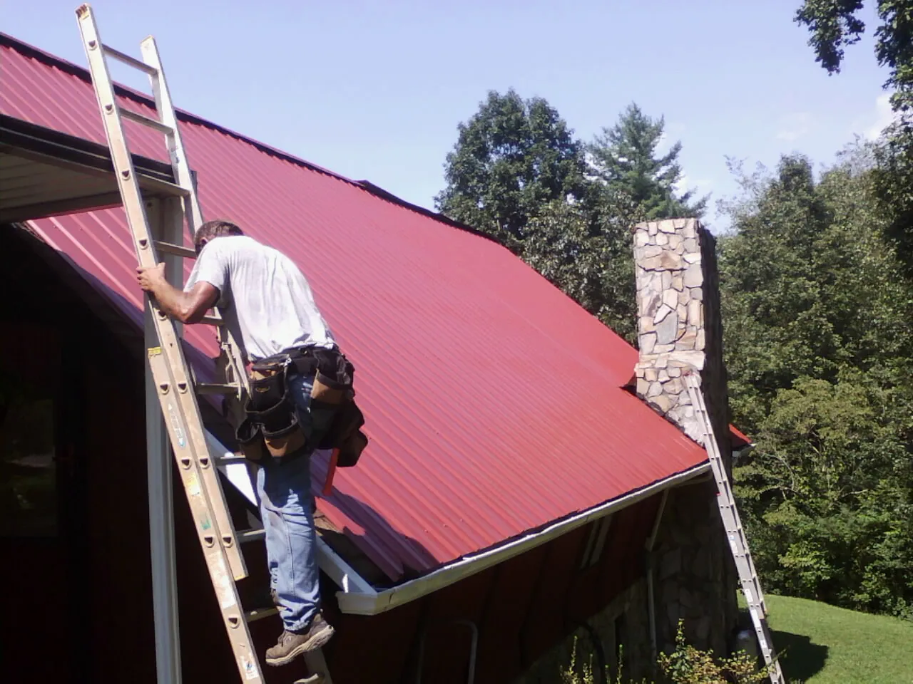 Roofer on a ladder beside a red metal roof and stone chimney — Soffit & Fascia Repair by Roofing Brothers in Grand Prairie